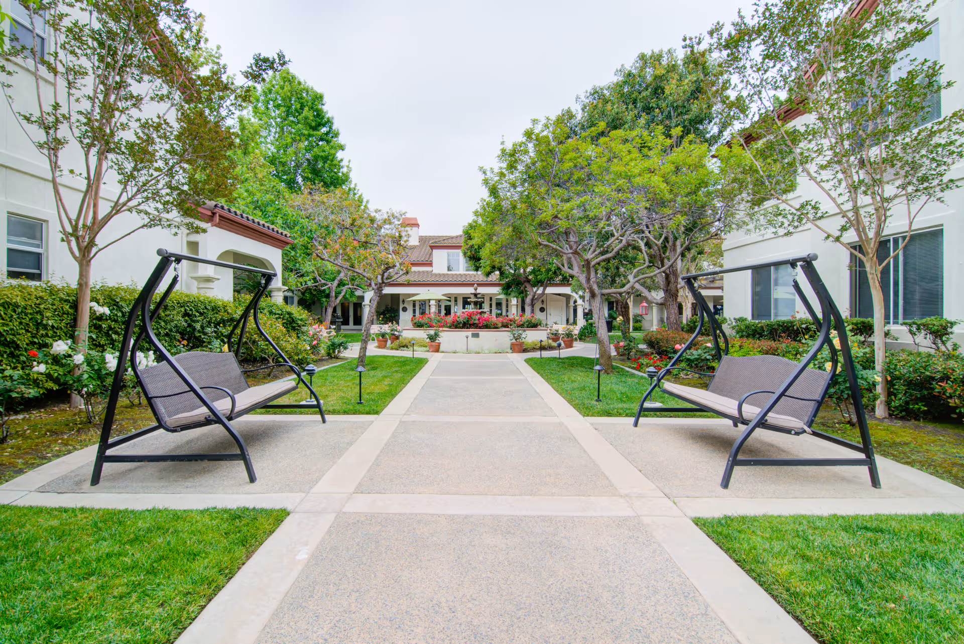 Outdoor courtyard area at Aegis Living Shadowridge featuring two metal-framed swing benches facing each other on a paved walkway, surrounded by green grass, trees, and shrubs with a building in the background.