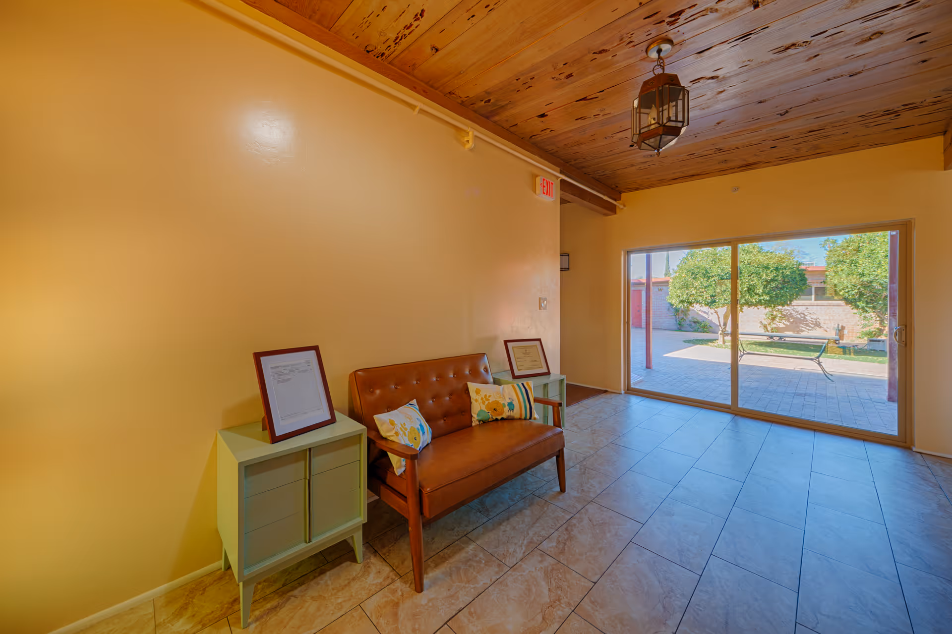 A small seating area with a brown leather loveseat adorned with two decorative pillows, flanked by two small green side tables each holding a framed document. The room has yellow walls, a wooden ceiling with a hanging lantern-style light fixture, and large sliding glass doors that open to an outdoor patio with trees and a bench.