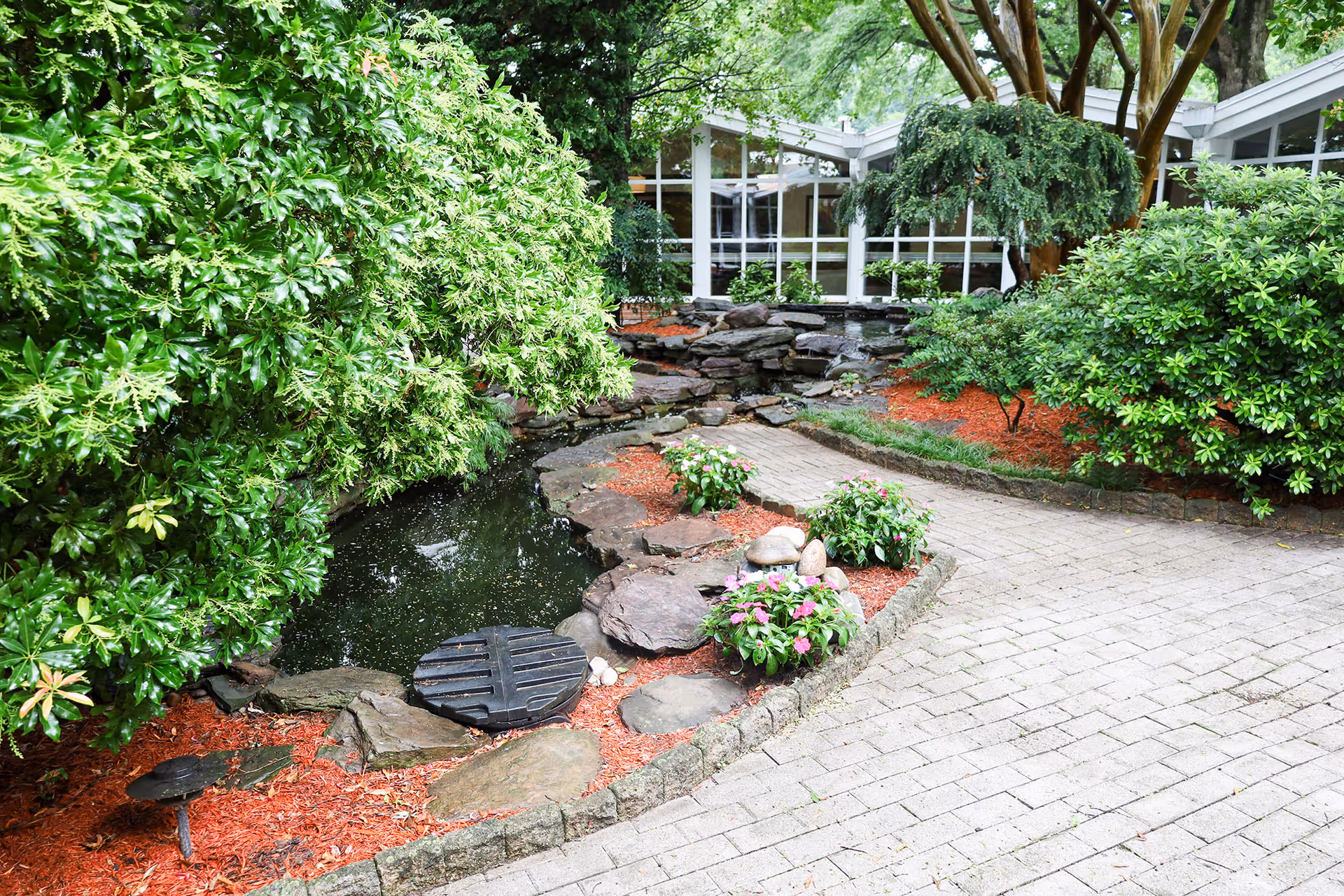 A serene outdoor garden area at Annandale Healthcare Center featuring a small pond surrounded by rocks, lush green bushes, flowering plants, and a paved walkway. The background shows large windows of the building with trees providing shade.