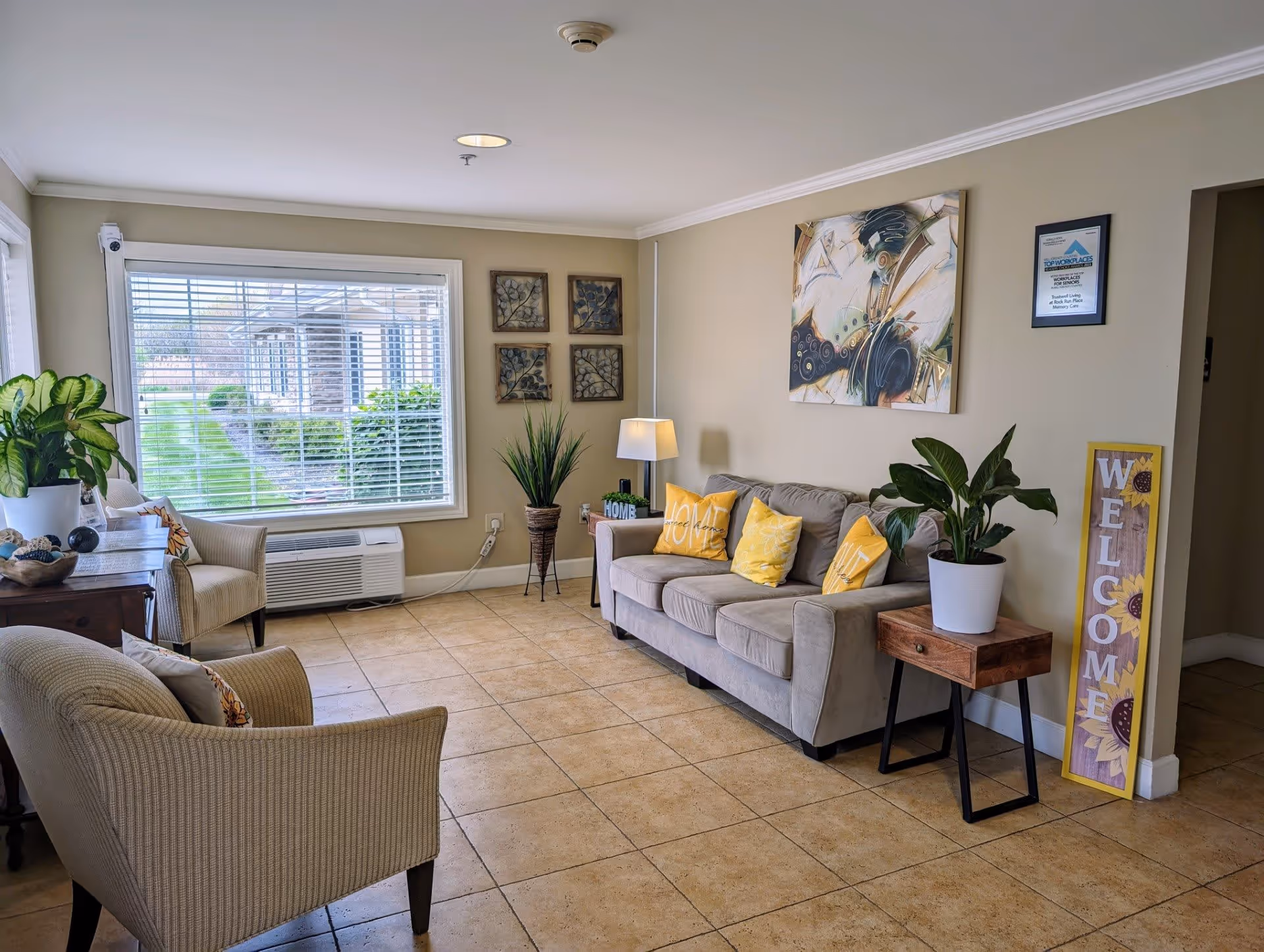 A cozy living room area with a beige sofa adorned with yellow and patterned pillows, two beige armchairs, a wooden side table with a potted plant, a floor lamp, and decorative wall art. A large window with white blinds lets in natural light, and a welcome sign with sunflowers leans against the wall near an open doorway.