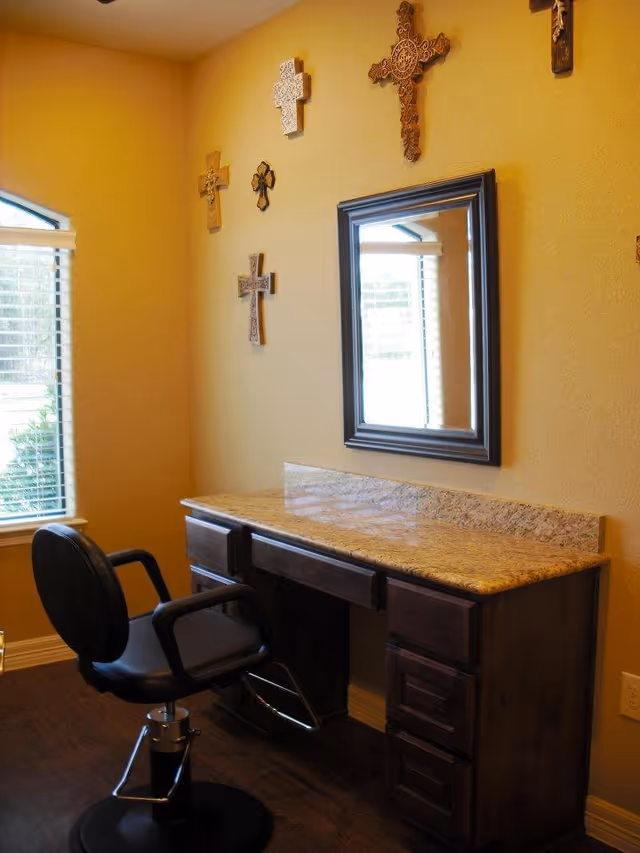 A small room with a wooden vanity desk topped with a granite surface, a black swivel chair in front of it, a rectangular mirror mounted on the wall above the desk, and several decorative crosses hanging on the wall. A window with blinds is visible on the left side, letting in natural light.