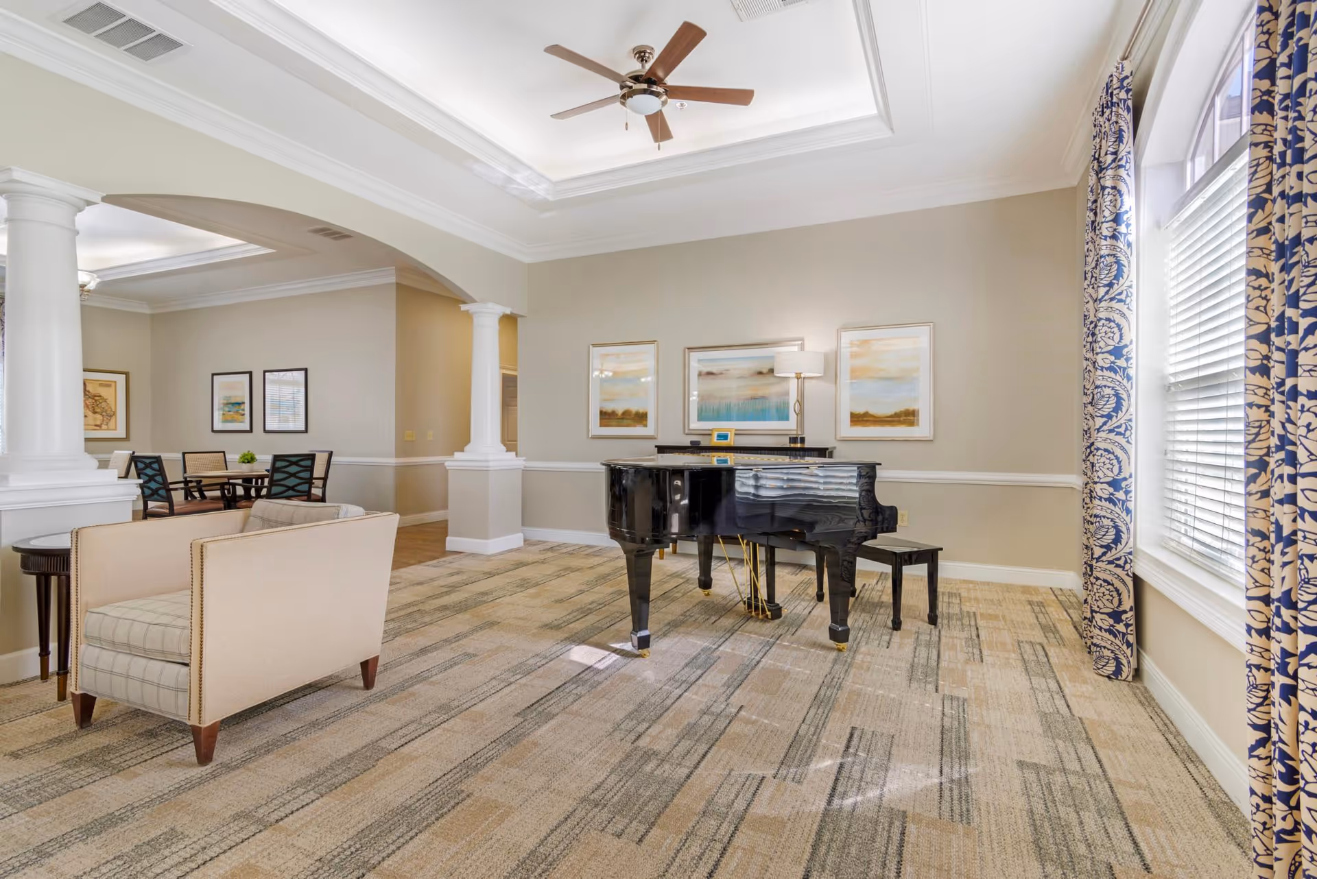 A spacious, well-lit room with a black grand piano and matching bench in the center. The room features beige walls with white trim, three framed landscape paintings above the piano, and a floor lamp. To the left, there is a beige armchair with plaid upholstery and a small round side table. In the background, a dining area with a table and chairs is visible. Large windows with blue and white patterned curtains allow natural light to fill the room. The ceiling has a tray design with a ceiling fan.