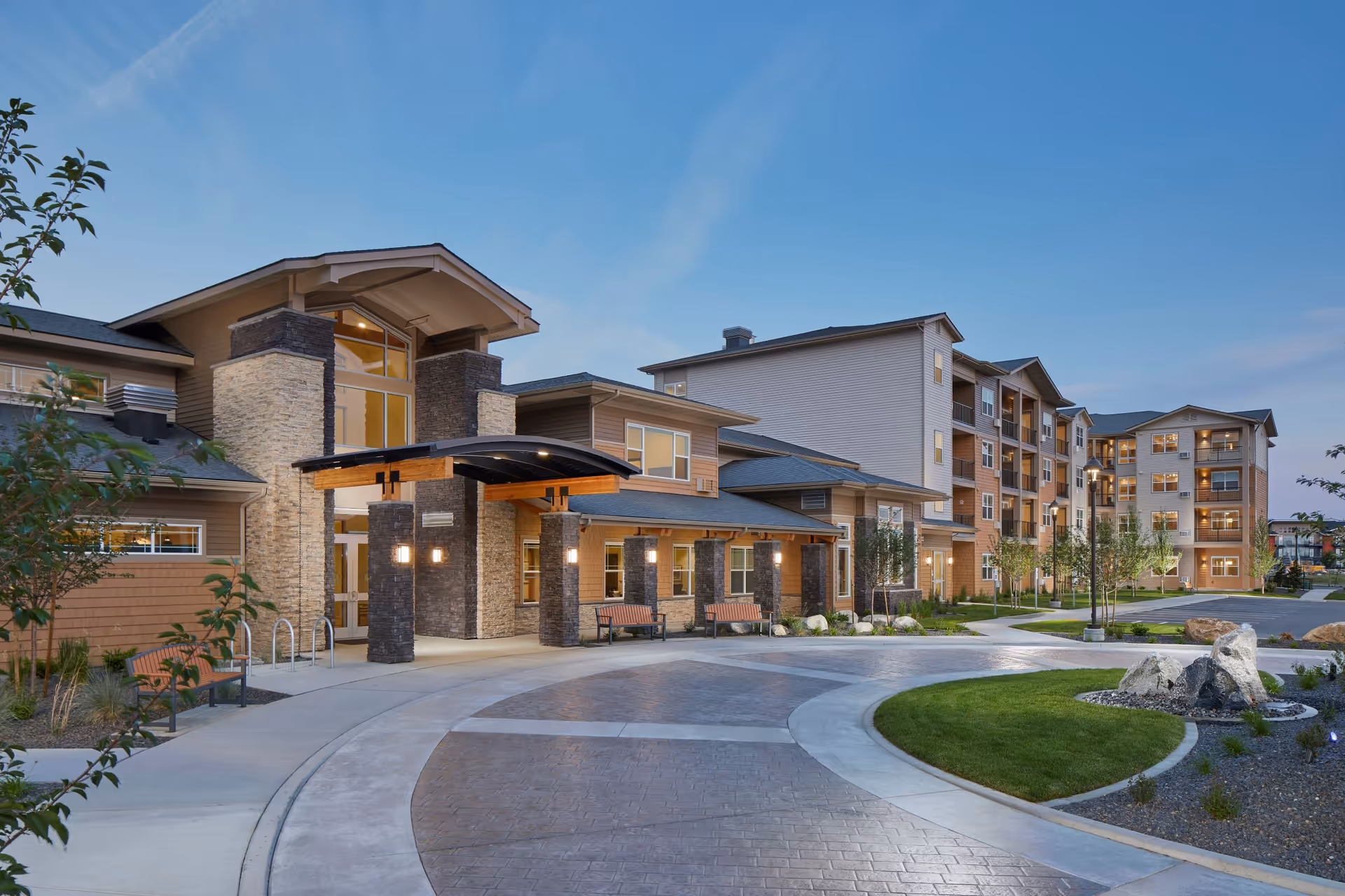 Exterior view of a modern senior living facility building at dusk with well-maintained landscaping, a curved driveway, benches, and multiple windows illuminated from inside.