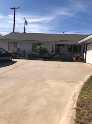 Exterior view of a single-story building with a gray roof and white walls, surrounded by a concrete driveway and some greenery. There are potted plants near the entrance and a parked vehicle on the left side under a clear blue sky.