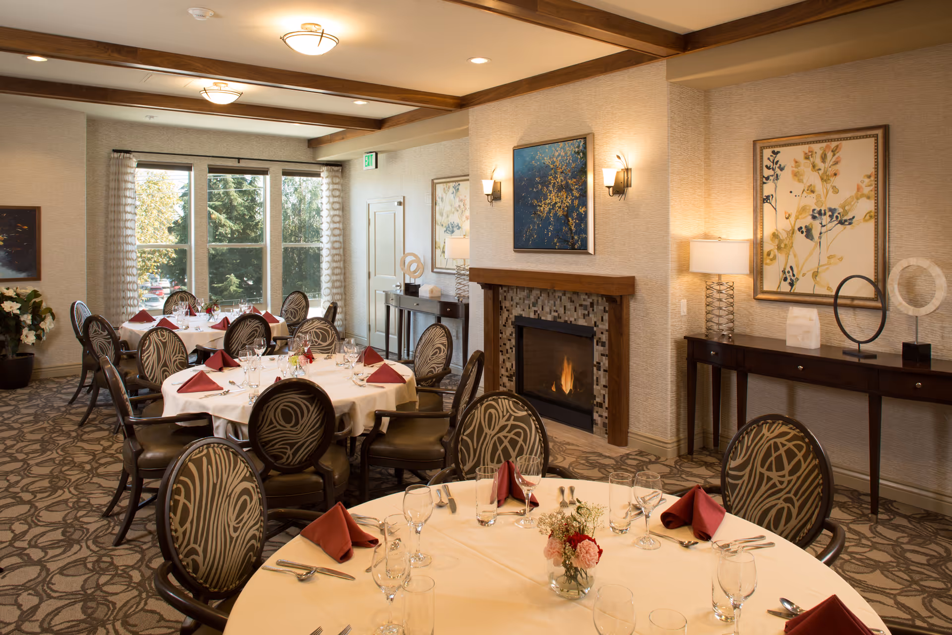 Dining room with round tables set for a meal, upholstered chairs, a lit fireplace, and wall artwork.