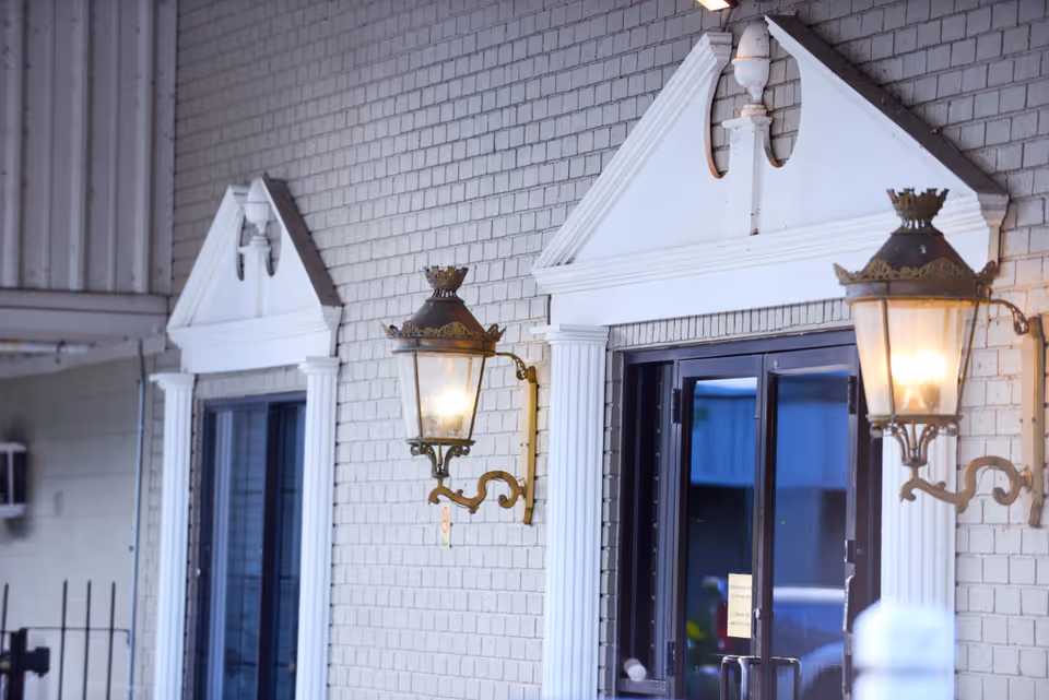 Exterior view of a building wall with two decorative wall-mounted lanterns lit on either side of a glass door entrance framed by white architectural trim and columns.