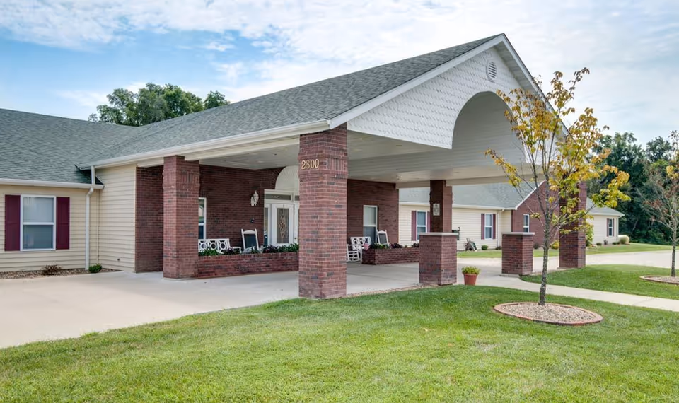 Exterior view of Victorian Place of Washington Senior Living showing a covered entrance supported by brick pillars, with a driveway and well-maintained lawn and small trees in front.