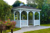 A white wooden gazebo with a shingled roof situated on a paved path surrounded by green grass and trees. There is a planter with pink flowers on the left side of the image.