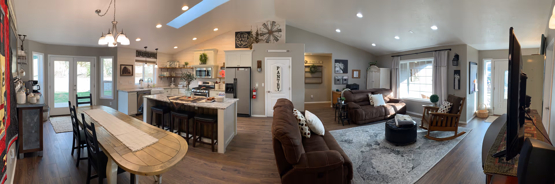 Panoramic view of an open-concept living space in a residential care facility. The image shows a kitchen with white cabinets, a granite countertop island with bar stools, and a dining area with a wooden table and chairs. Adjacent to the kitchen is a living room with brown sofas, a rocking chair, a large window with curtains, and a TV on a stand. The space has wood flooring, recessed lighting, and a door labeled 'PANTRY'.
