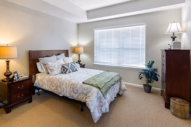 A well-lit bedroom with a large window covered by white blinds. The room features a bed with a wooden headboard, patterned bedding, multiple pillows, and a green throw blanket. There are two wooden nightstands with lamps on either side of the bed, a wooden dresser with a lamp and decorative items, a potted plant near the window, and a woven basket on the floor.