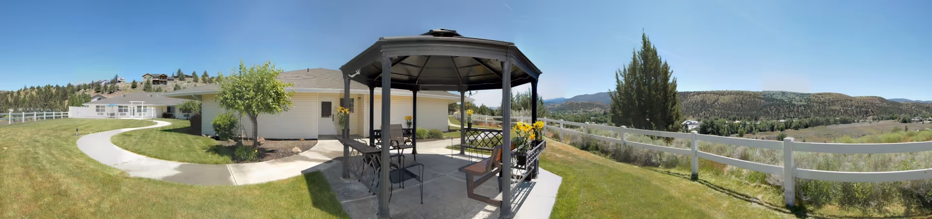Outdoor area of a senior living facility featuring a gazebo with seating, a paved walkway, a white fence, green grass, trees, and a scenic view of hills and mountains in the background under a clear blue sky.