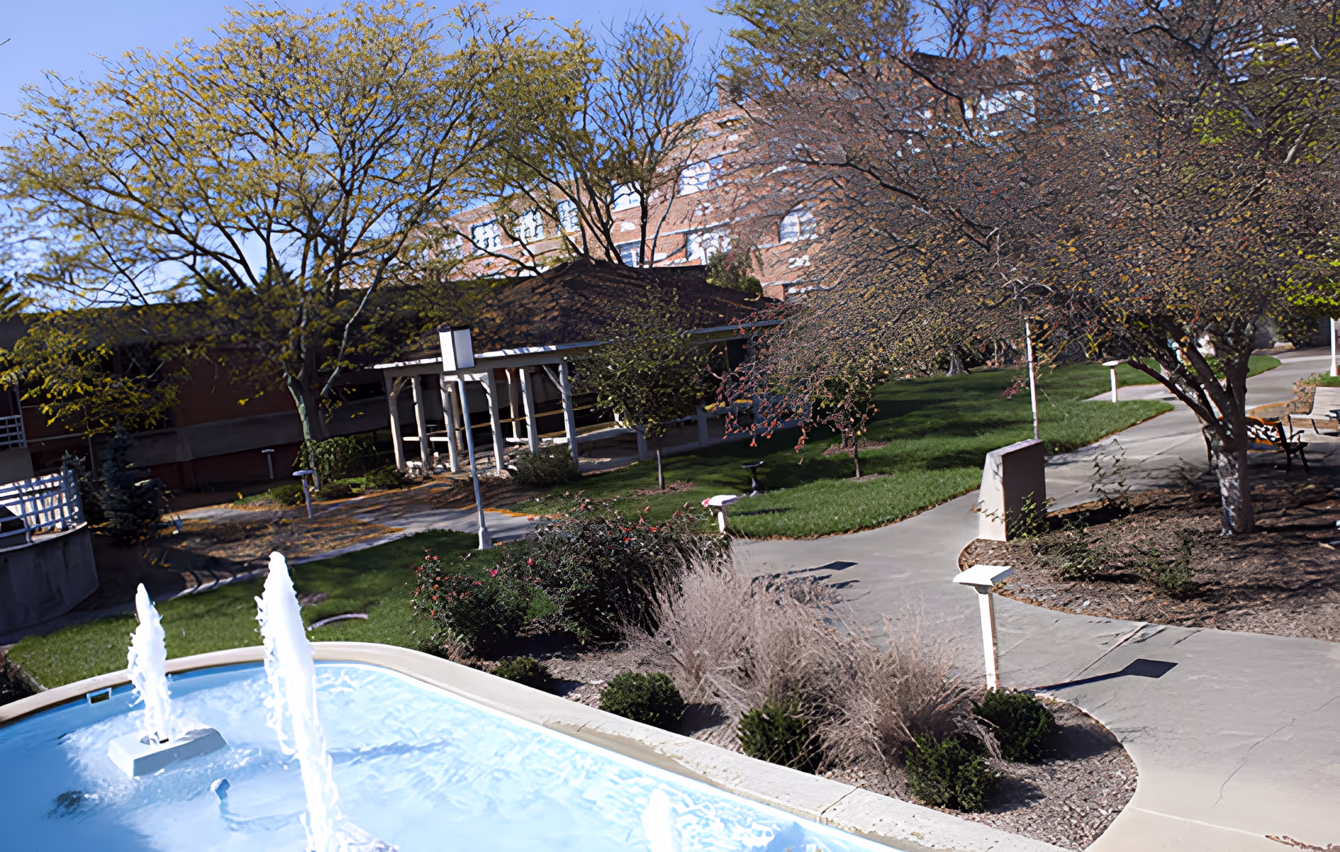 Outdoor garden area at Indiana Masonic Home Community Center featuring a water fountain with three jets, paved walkways, trees with sparse foliage, bushes, and a building in the background under a clear blue sky.