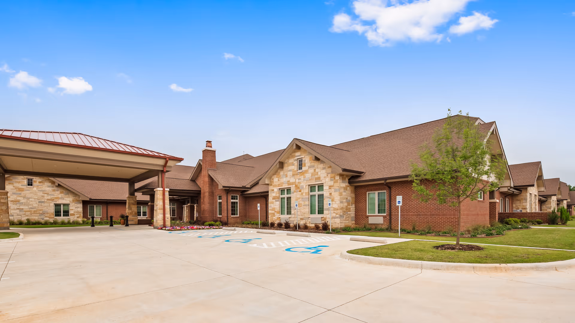 Exterior view of Legend of Mansfield senior living facility showing a single-story building with a combination of brick and stone facade, a covered entrance, a driveway with handicap parking spaces, and a small landscaped area with a tree and grass under a clear blue sky.