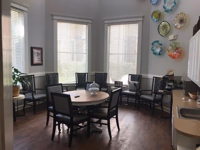 A bright room with three large windows covered by blinds, featuring a round wooden table surrounded by six chairs in the center. Additional chairs line the walls, and there is a small kitchenette area with a sink and countertop on the right. Decorative glass wall art is displayed on the upper right wall.