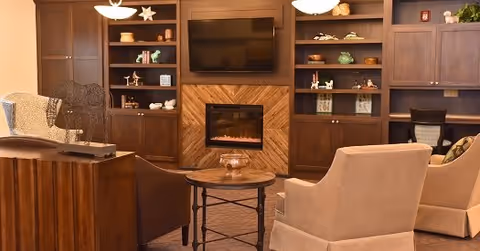 A cozy living room area with beige and brown armchairs arranged around a round wooden coffee table. The background features built-in wooden shelves with decorative items and a flat-screen TV mounted above a modern electric fireplace.