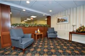 A waiting area with two blue patterned armchairs, a small wooden table with decorative items, a floral arrangement on a side table, and a large mirror on the wall reflecting the ceiling lights. The carpet has a dark floral pattern and the walls are decorated with striped wallpaper and a framed picture.