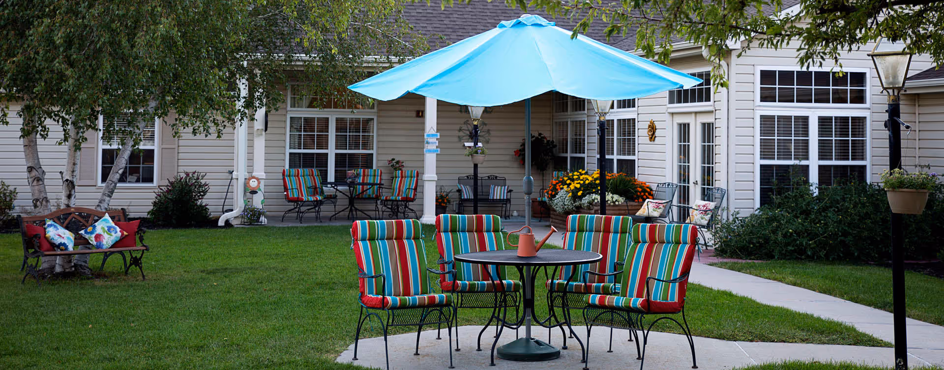 Outdoor patio area with a round table and six colorful striped cushioned chairs under a large blue umbrella. There is a watering can on the table. In the background, there are more chairs and tables on a covered porch attached to a beige building with large windows. The area is surrounded by green grass, trees, and flower beds.