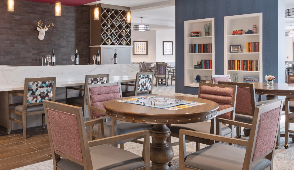 Communal lounge featuring a round game table surrounded by chairs, bookshelves, and a marble-topped serving counter with wine storage.