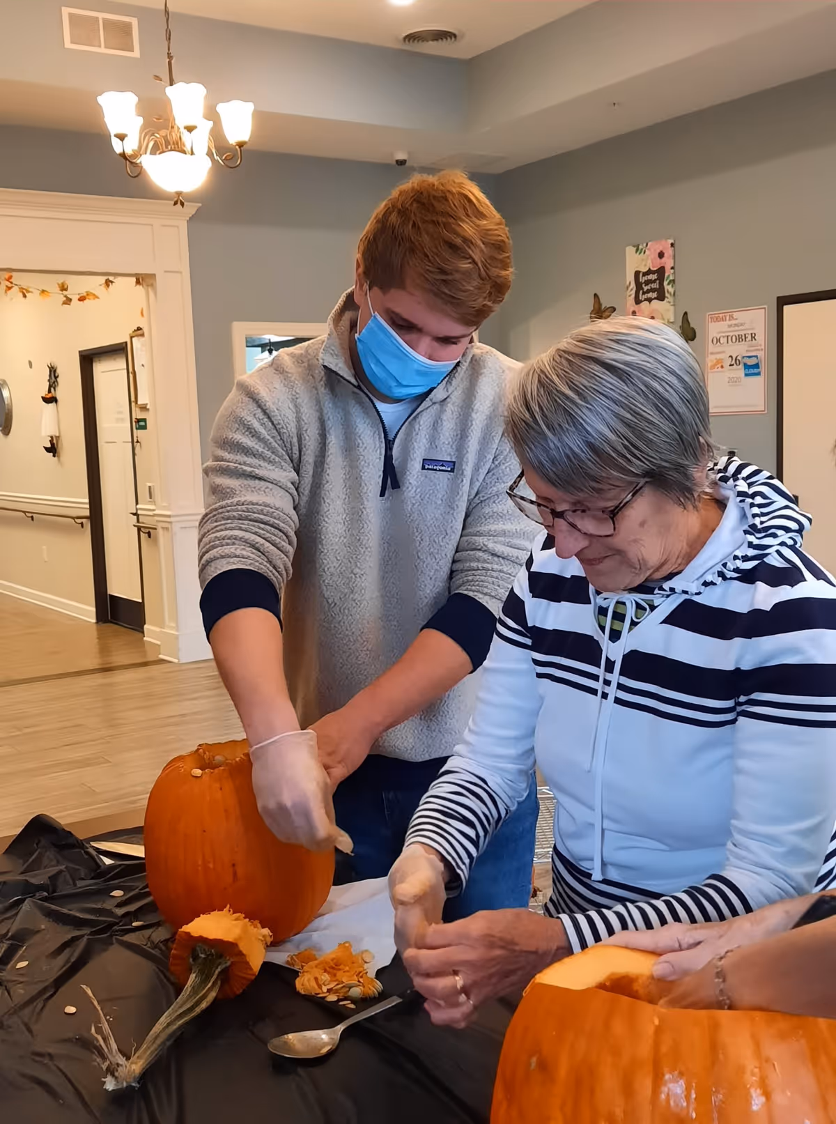 A young man wearing a face mask and gloves is helping an older woman with short gray hair and glasses carve a pumpkin indoors. They are seated at a table covered with a black plastic tablecloth, with pumpkin seeds and pulp visible on the table. The room has light blue walls, wooden flooring, and a chandelier hanging from the ceiling.