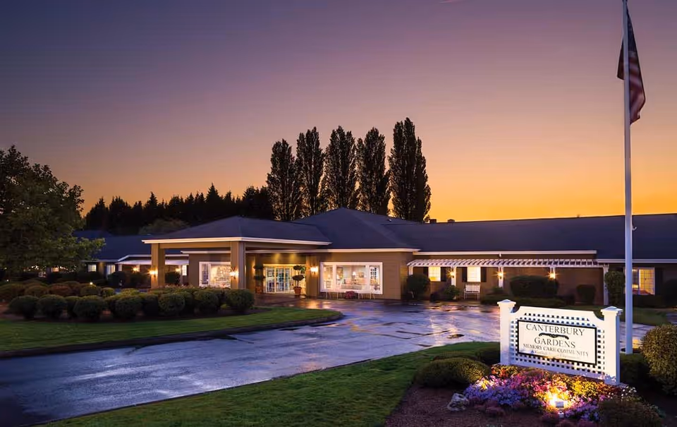 Exterior view of Canterbury Gardens Memory Care Community building at dusk with illuminated entrance, manicured bushes, and a sign in the foreground. The sky is a gradient of warm colors from the setting sun.