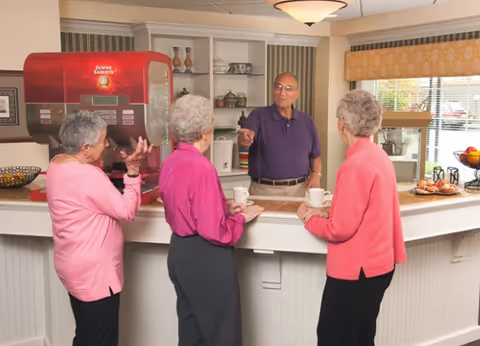 Three elderly women standing at a counter talking to an elderly man behind the counter in a cozy room with shelves, a large red coffee machine, and a popcorn machine near the window.