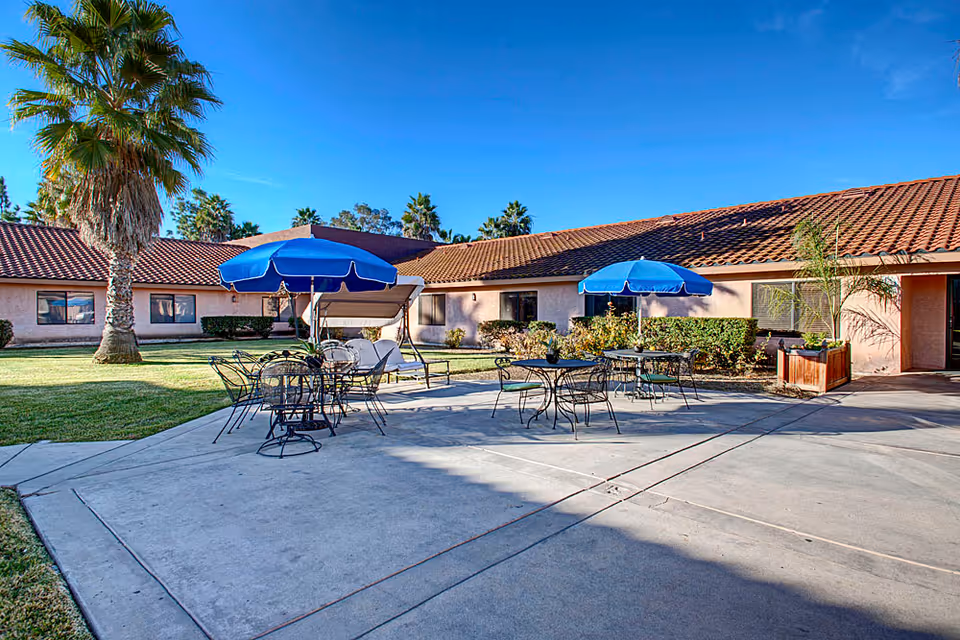 Outdoor patio area at Santa Fe Post Acute with metal tables and chairs, blue umbrellas providing shade, a swing bench, palm trees, and a building with a tiled roof in the background under a clear blue sky.