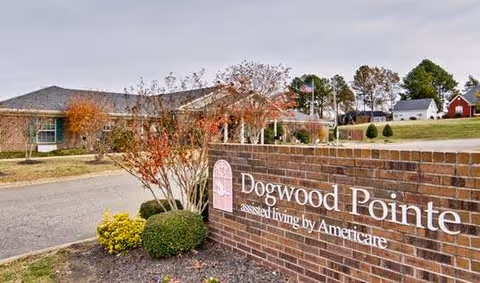 Exterior view of Dogwood Pointe assisted living facility showing a brick sign with the facility name and a building with trees and landscaping in the background under a cloudy sky.