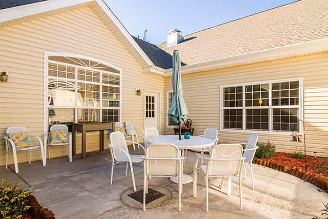 Outdoor patio area with a round table surrounded by eight white chairs and a closed green umbrella in the center. The patio is adjacent to a beige building with large windows and a door. There are some plants and mulch along the edges of the patio.