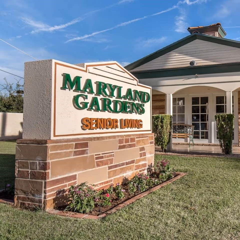 Outdoor view of the Maryland Gardens Senior Living facility sign with a building entrance in the background, surrounded by green grass and small plants under a blue sky.