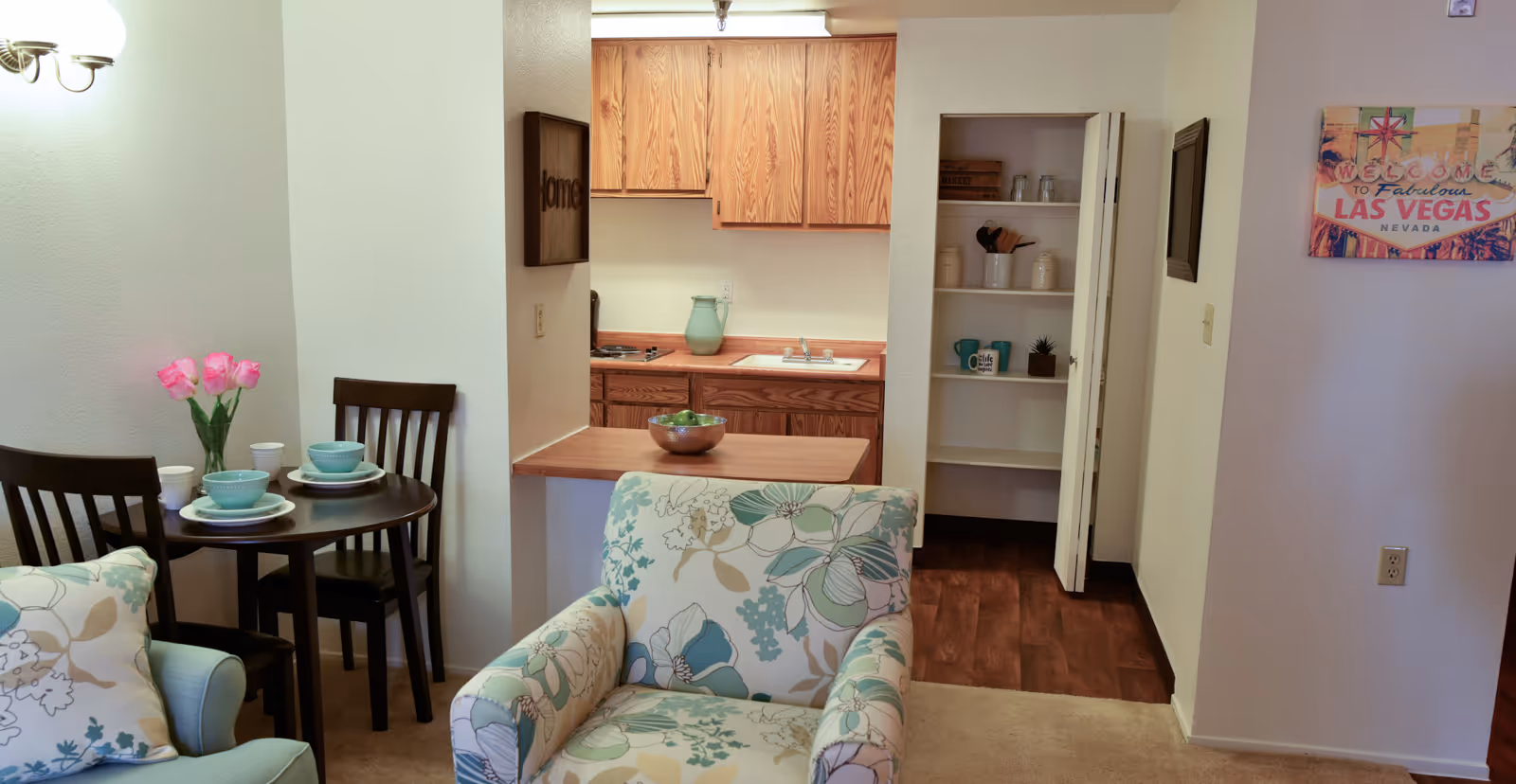 Open-plan living area with floral armchairs, a small dining table set for two, and a kitchenette with wooden cabinets.