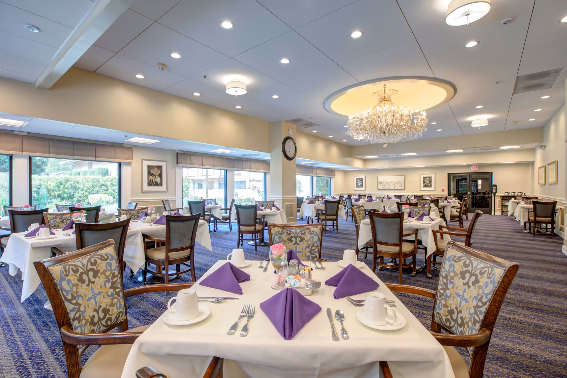 A spacious dining room in a senior living facility with multiple tables covered in white tablecloths, each set with purple folded napkins, white cups, and silverware. The room features large windows letting in natural light, patterned chairs, a blue carpet, and a large chandelier hanging from the ceiling.