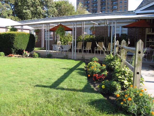 Outdoor patio area with a covered seating space featuring tables, chairs, and red umbrellas. The patio is adjacent to a brick building and surrounded by a well-maintained lawn and flower beds with various colorful flowers and green plants. A wooden fence runs along one side of the patio.