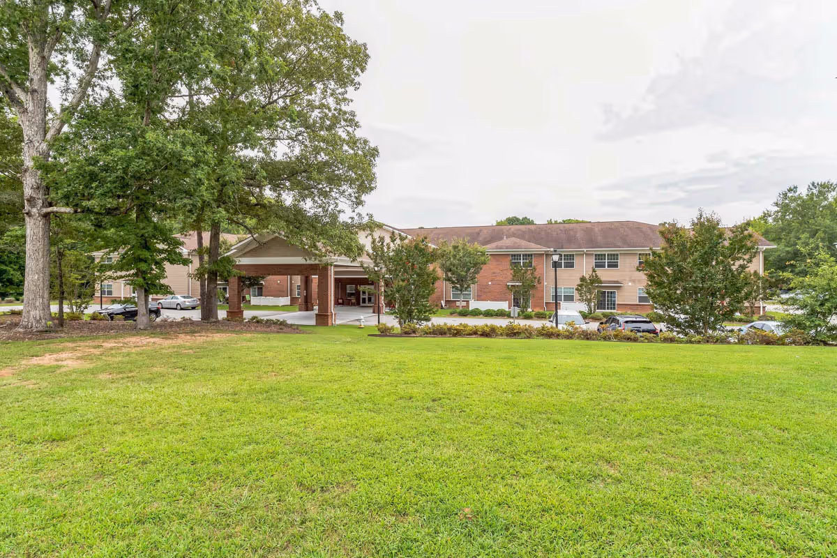 Exterior view of a senior living facility building with a covered entrance, surrounded by green grass, trees, and parked cars under a cloudy sky.