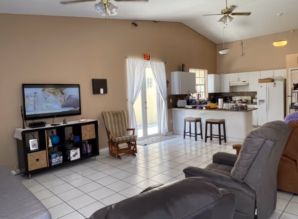 A bright living area with beige walls and white tiled floor featuring a mounted flat-screen TV above a black shelving unit with various items. There is a striped rocking chair near glass doors with white curtains leading outside. The open kitchen has white cabinets, a refrigerator, stove, and a counter with two stools. Comfortable recliner chairs and a brown sofa are partially visible in the foreground.