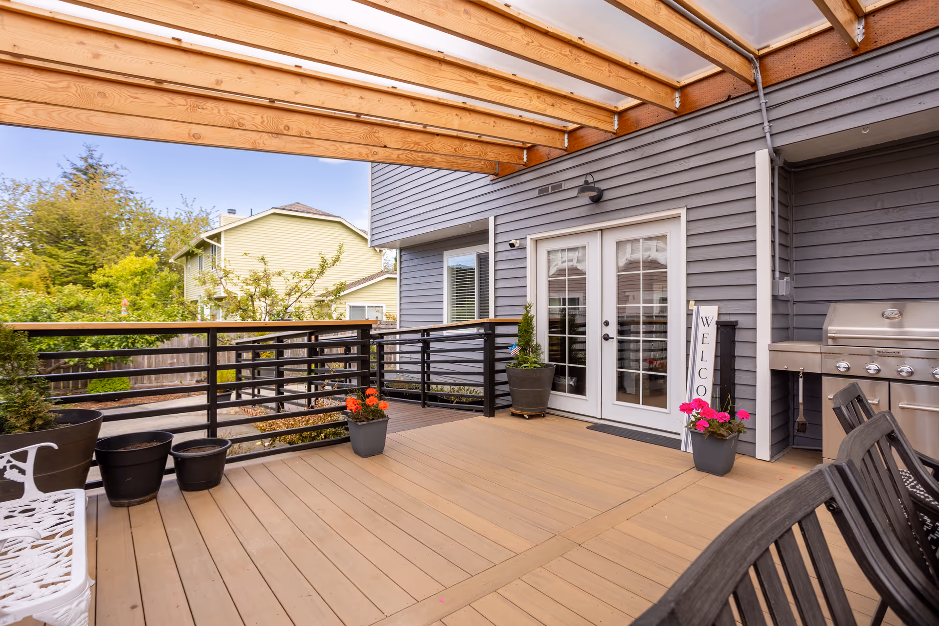A covered wooden deck with potted plants, outdoor seating, a grill, and French doors leading into a gray house.