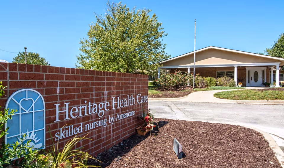 Brick sign reading 'Heritage Health Care' at the landscaped driveway entrance to a single-story skilled nursing facility.