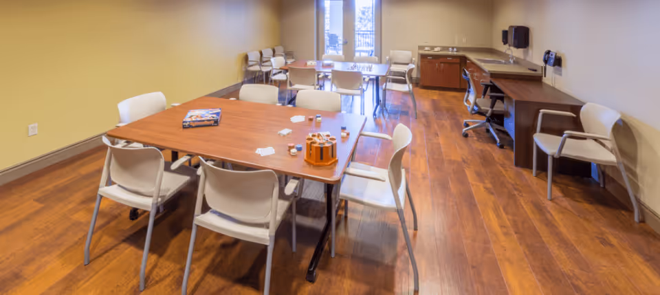 A well-lit room with wooden flooring featuring a large rectangular table surrounded by white chairs. On the table are some board games and small containers. In the background, there are additional chairs and a desk with office supplies against the wall. A glass door leads to an outdoor area.