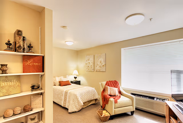 A cozy senior living bedroom with a neatly made bed featuring beige and white bedding, a bedside table with a lamp, a comfortable armchair draped with a red throw blanket and a pillow, a basket on the floor, a large window with blinds, and a TV on a wooden stand. A bookshelf with decorative items and framed art is visible on the left side of the image.