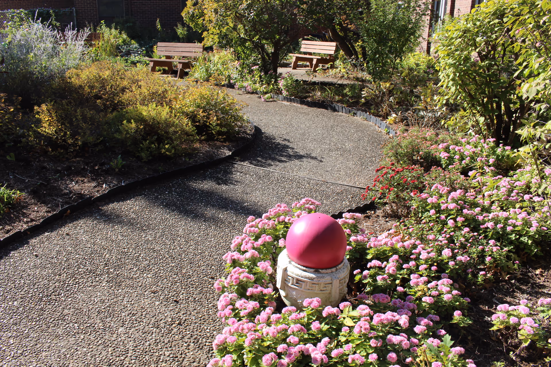A garden pathway winding through a landscaped area with various green shrubs and pink flowers. There are two wooden benches in the background and a decorative stone pedestal with a pink spherical ornament in the foreground.