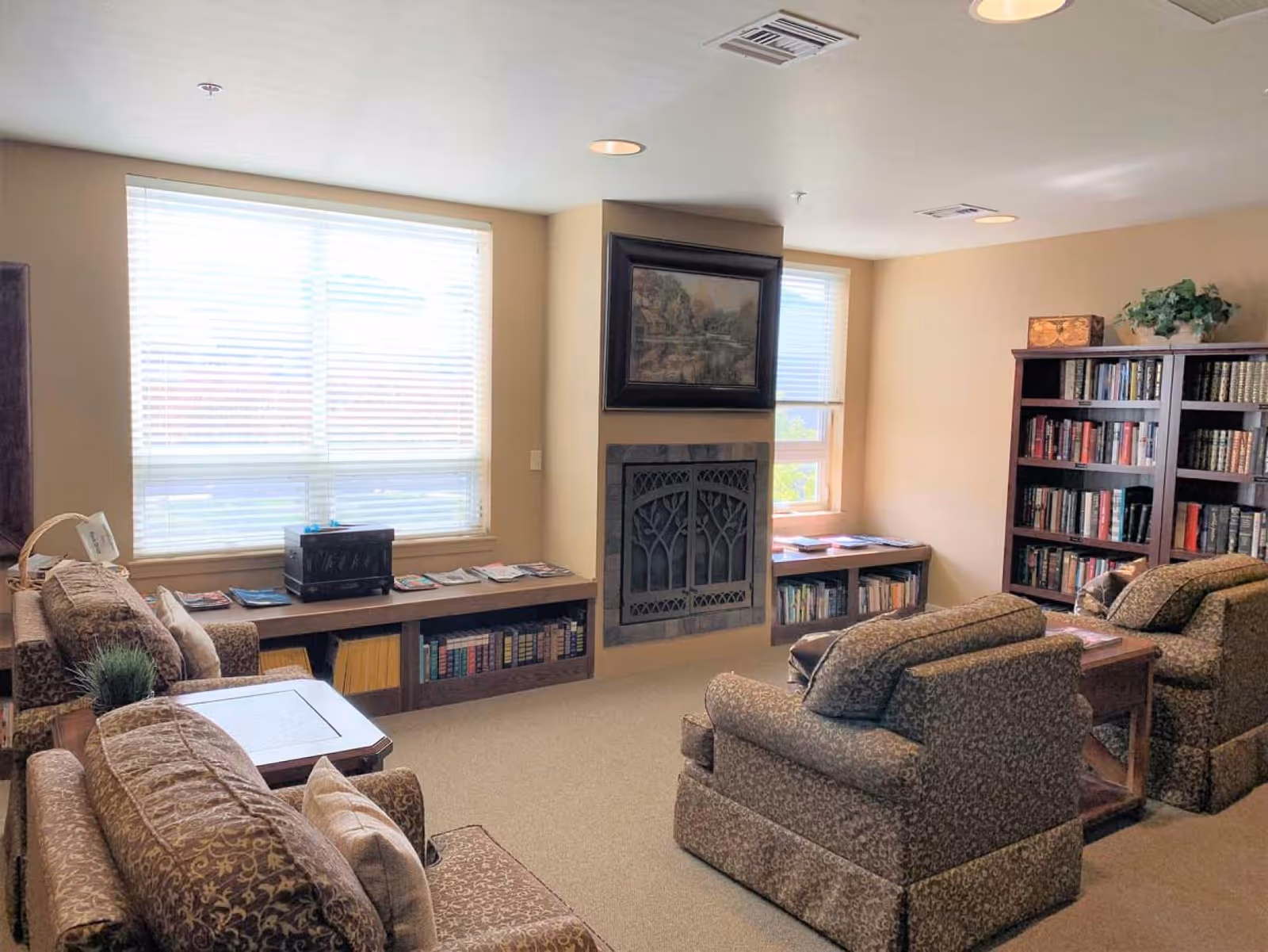 Comfortable common living room with upholstered armchairs, bookshelves, large windows, and a decorative fireplace.