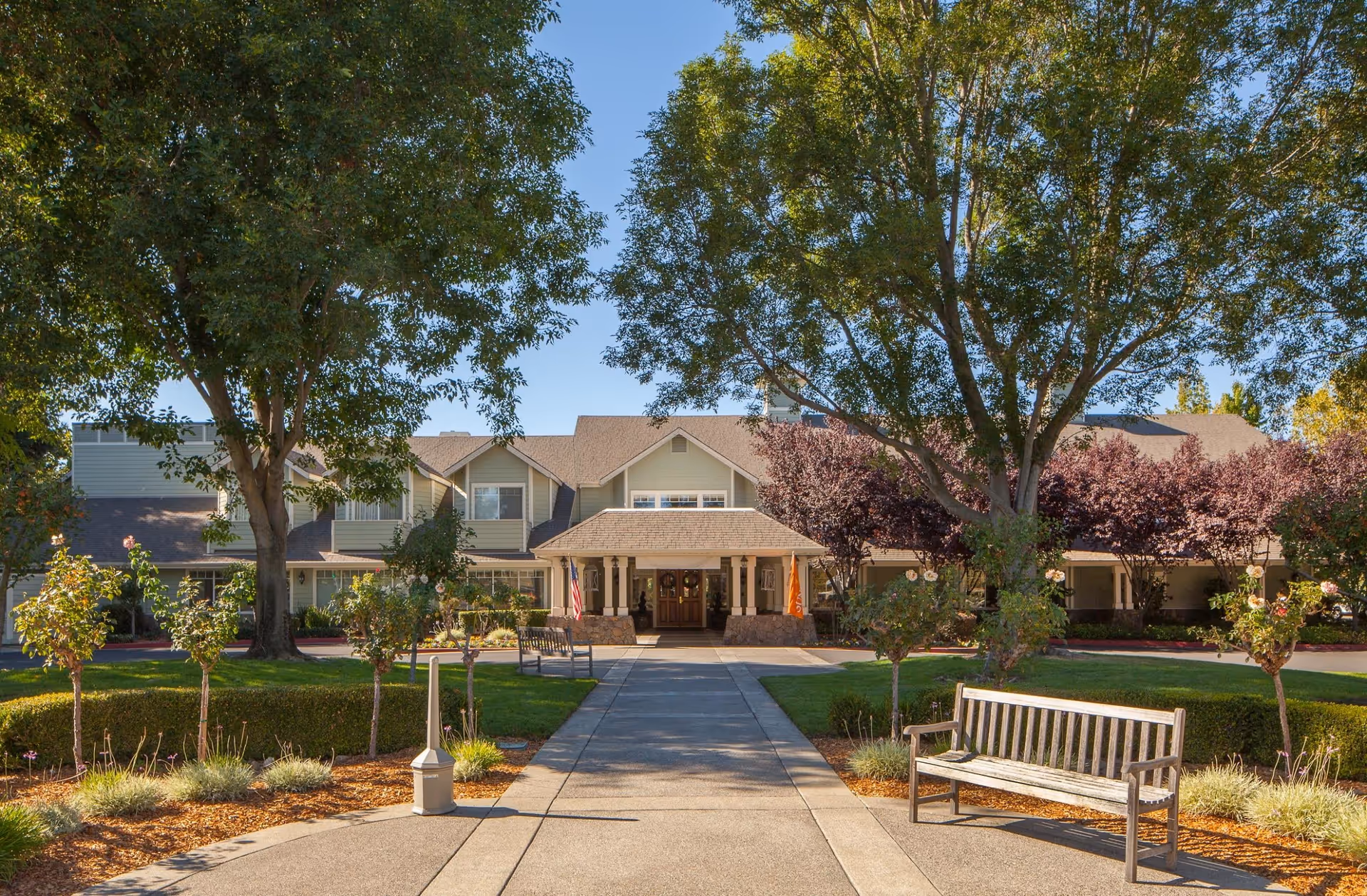 Front exterior view of Windsong of Sonoma Senior Living facility with a paved walkway leading to the entrance, surrounded by trees, bushes, and benches on a sunny day.
