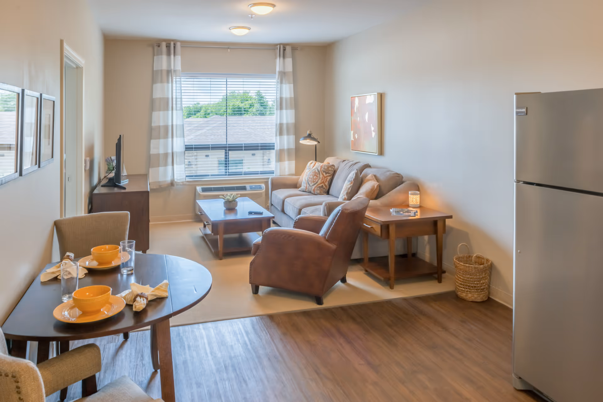 A cozy senior living apartment featuring a small dining table set with two place settings, a comfortable living area with a sofa, armchair, coffee table, side table with a lamp, and a large window with striped curtains. A refrigerator is visible on the right side, and the room has a mix of carpet and wood flooring.