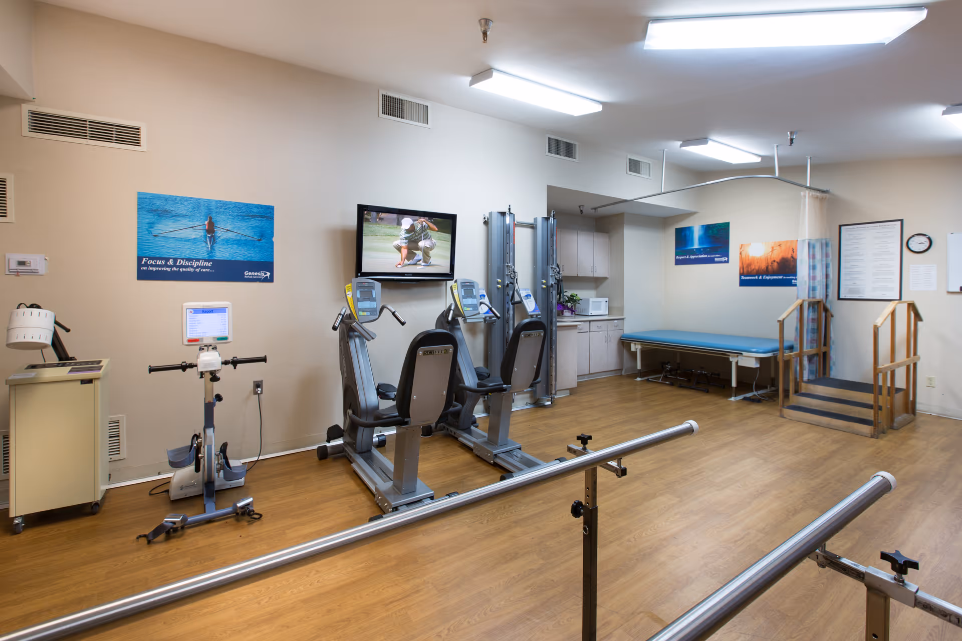 Physical therapy room with exercise machines, parallel bars, a treatment table and a small practice staircase.
