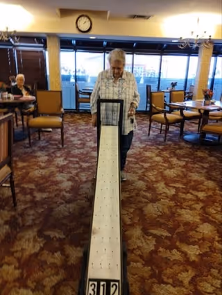 An elderly woman playing a tabletop shuffleboard game in a common area with carpeted floor and several chairs and tables. Another elderly person is seated in the background near large windows letting in natural light.