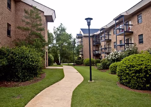 A paved walkway curves through a landscaped outdoor area between two multi-story brick buildings with balconies. The area is surrounded by green grass, bushes, and trees, with a lamppost along the path.