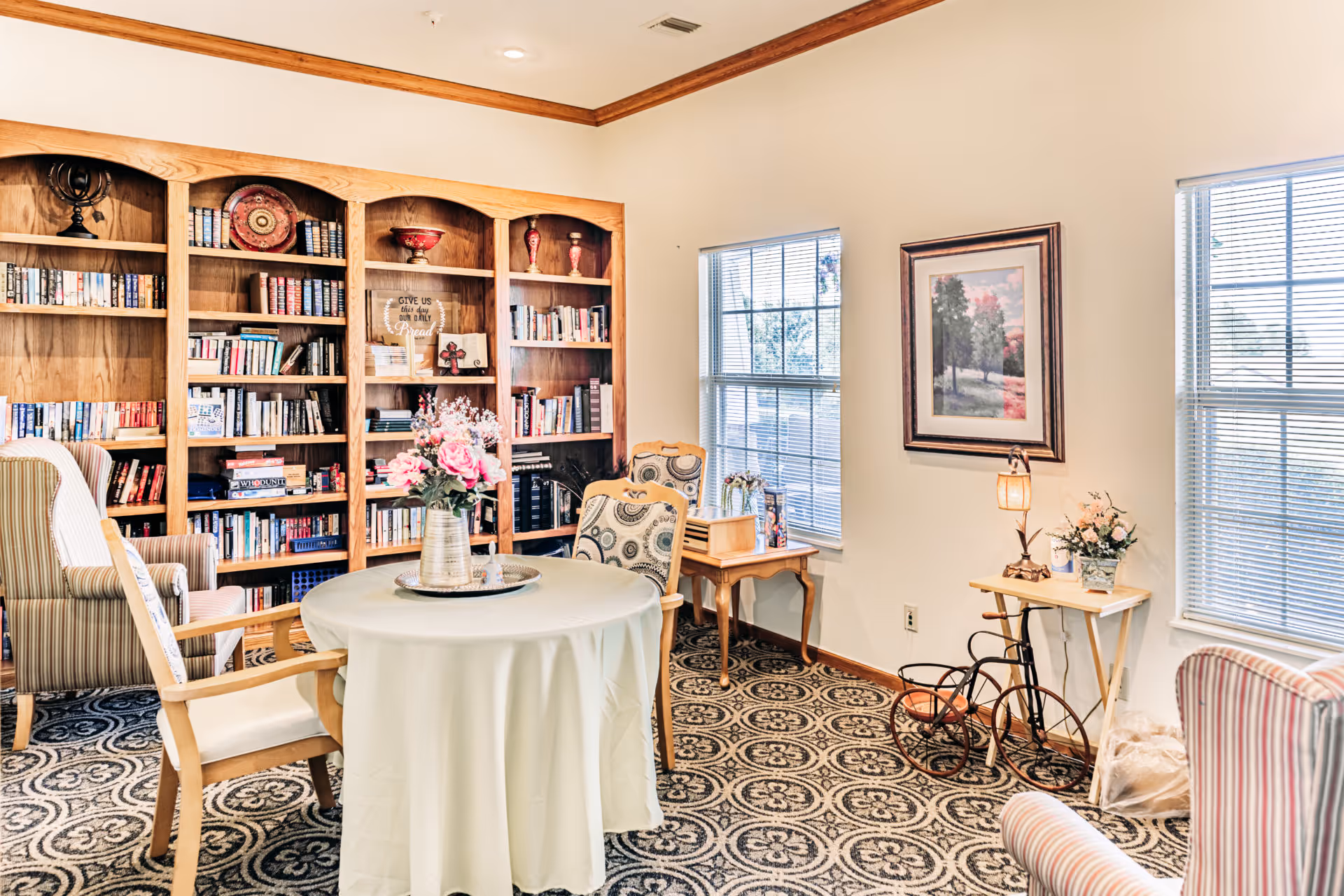 Bright common room with a round table, armchairs, patterned carpet, and a large wooden bookshelf filled with books.