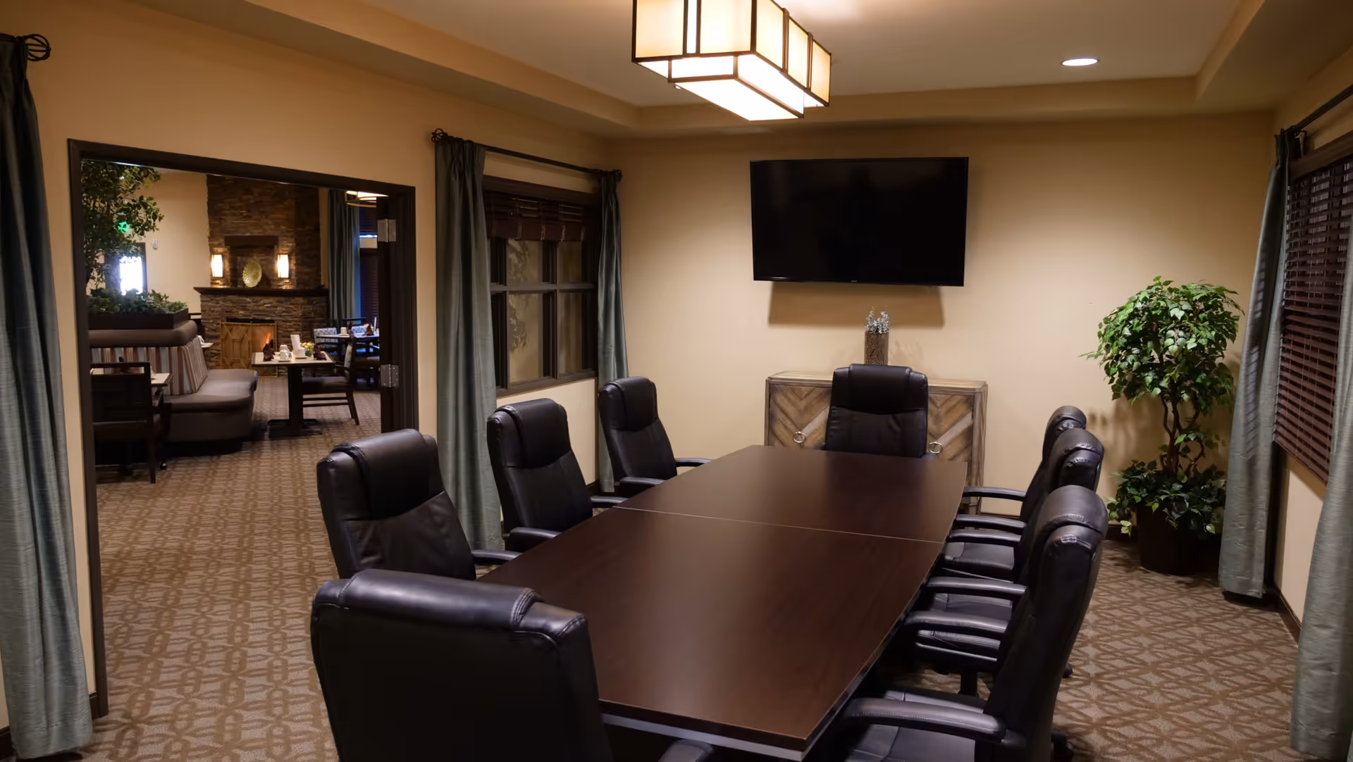 A conference room with a long dark wood table surrounded by eight black leather chairs. The room has beige walls, carpeted floor with a patterned design, two windows with green curtains, a flat-screen TV mounted on the wall, a wooden cabinet beneath the TV, and a potted plant in the corner. Through an open doorway, a cozy seating area with a stone fireplace and dining tables is visible.