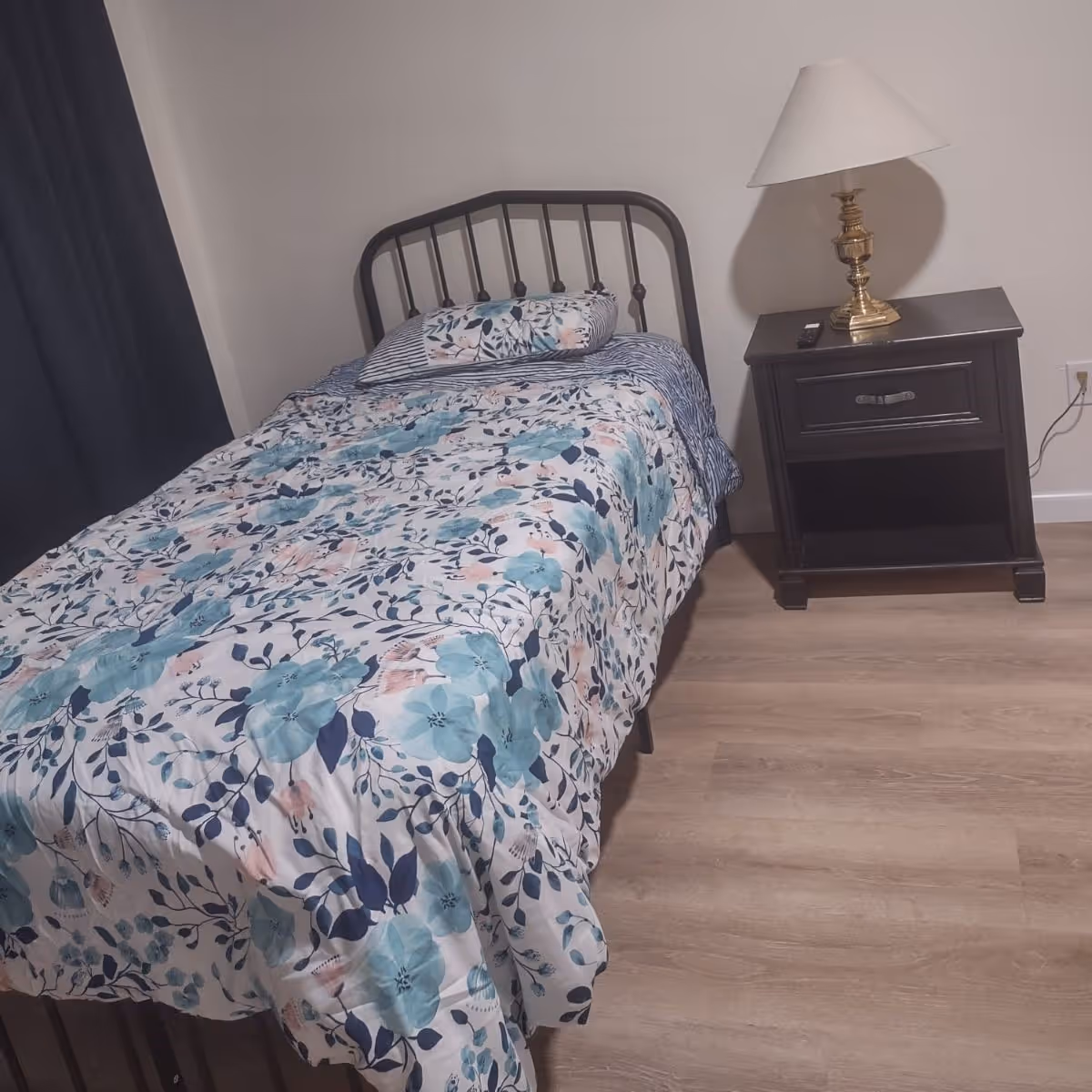 A small bedroom with a single metal bed frame, a floral patterned bedspread in shades of blue and pink, and a matching pillow. Next to the bed is a dark wooden nightstand with a drawer and an open shelf, topped with a brass lamp with a white lampshade. The floor is light wood, and there is a dark curtain on the left side of the image.