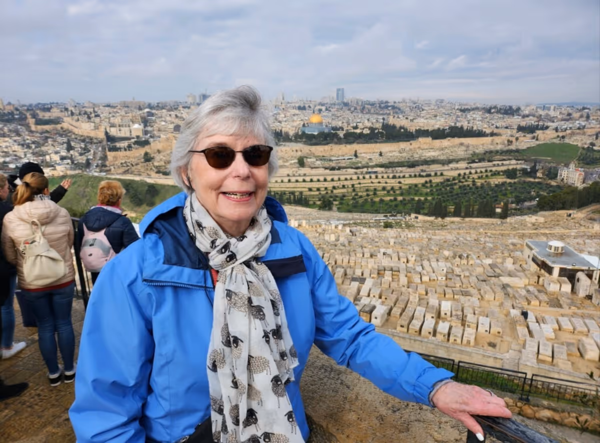 An elderly woman wearing sunglasses, a blue jacket, and a scarf with sheep patterns stands outdoors with a scenic view of a historic city and a large cemetery in the background. Several other people are also visible in the background, looking at the view.