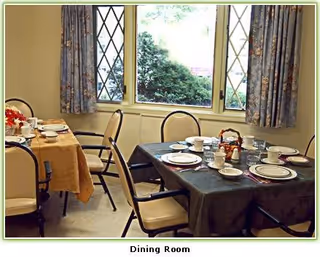 A dining room with two tables set for a meal. Each table has plates, cups, saucers, and cutlery arranged neatly. The tables are covered with tablecloths, one yellow and one dark green. There are several chairs around the tables. The room has windows with floral curtains and a view of greenery outside.